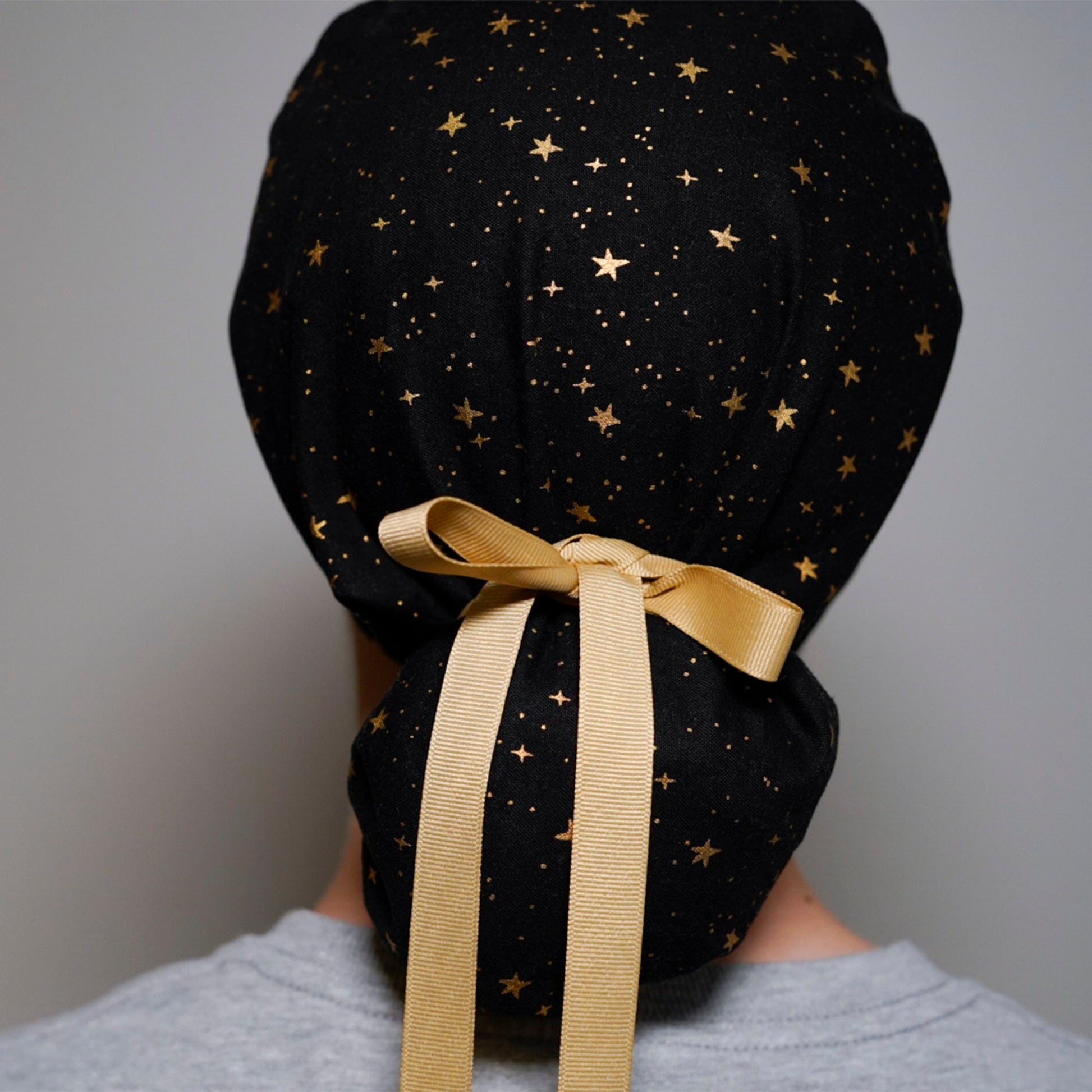 Black scrub hat with a gold star pattern and a gold satin ribbon tied in a bow at the back.