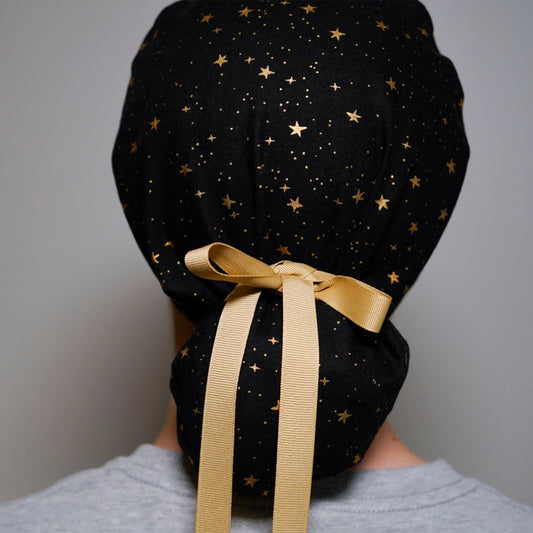 Black scrub hat with a gold star pattern and a gold satin ribbon tied in a bow at the back.