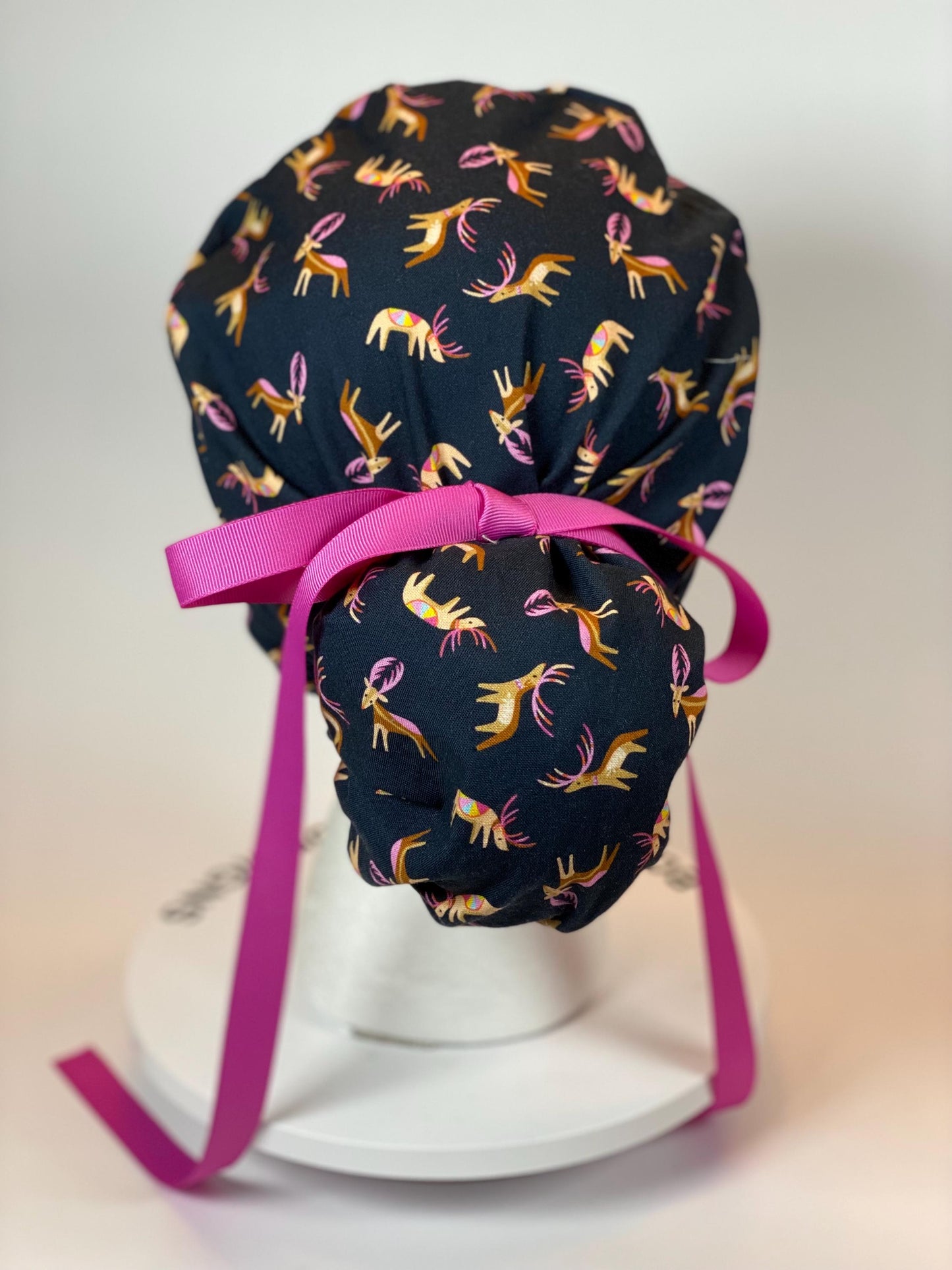 navy ponytail scrub cap with pouch that ties around hair bun. Cap has navy fabric with small brown reindeer with pink accents and a pink ribbon.