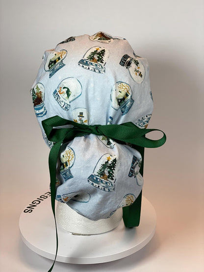 light blue fabric scrub cap with small snow globes featuring snow men, bears, and trees. The cap ties in a pouch around the hair with a green ribbon.