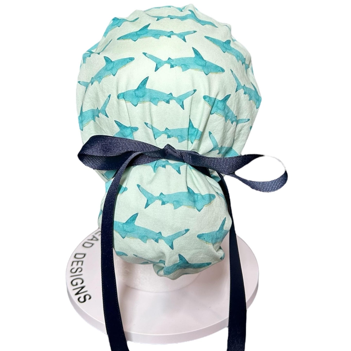 May include: A light green and white patterned scrub cap featuring watercolor-style blue sharks. The cap is tied with a navy blue ribbon. The cap is designed to be worn in a medical setting.