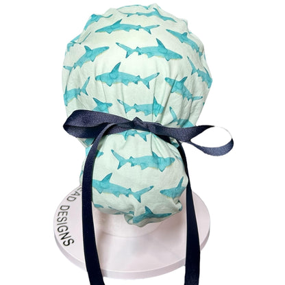 May include: A light green and white patterned scrub cap featuring watercolor-style blue sharks. The cap is tied with a navy blue ribbon. The cap is designed to be worn in a medical setting.