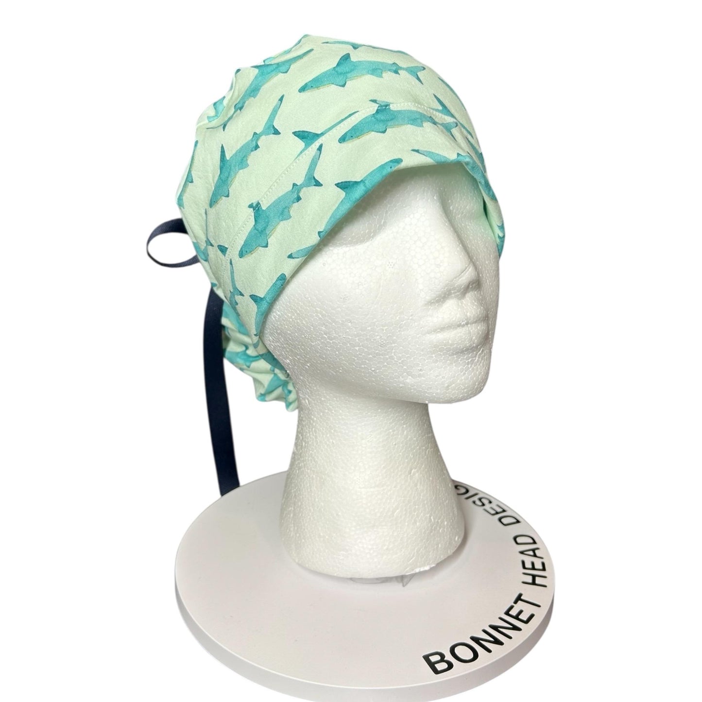 May include: A light green surgical scrub cap with a repeating pattern of blue watercolor sharks. The cap is tied with a navy blue ribbon. The cap is displayed on a white mannequin head.
