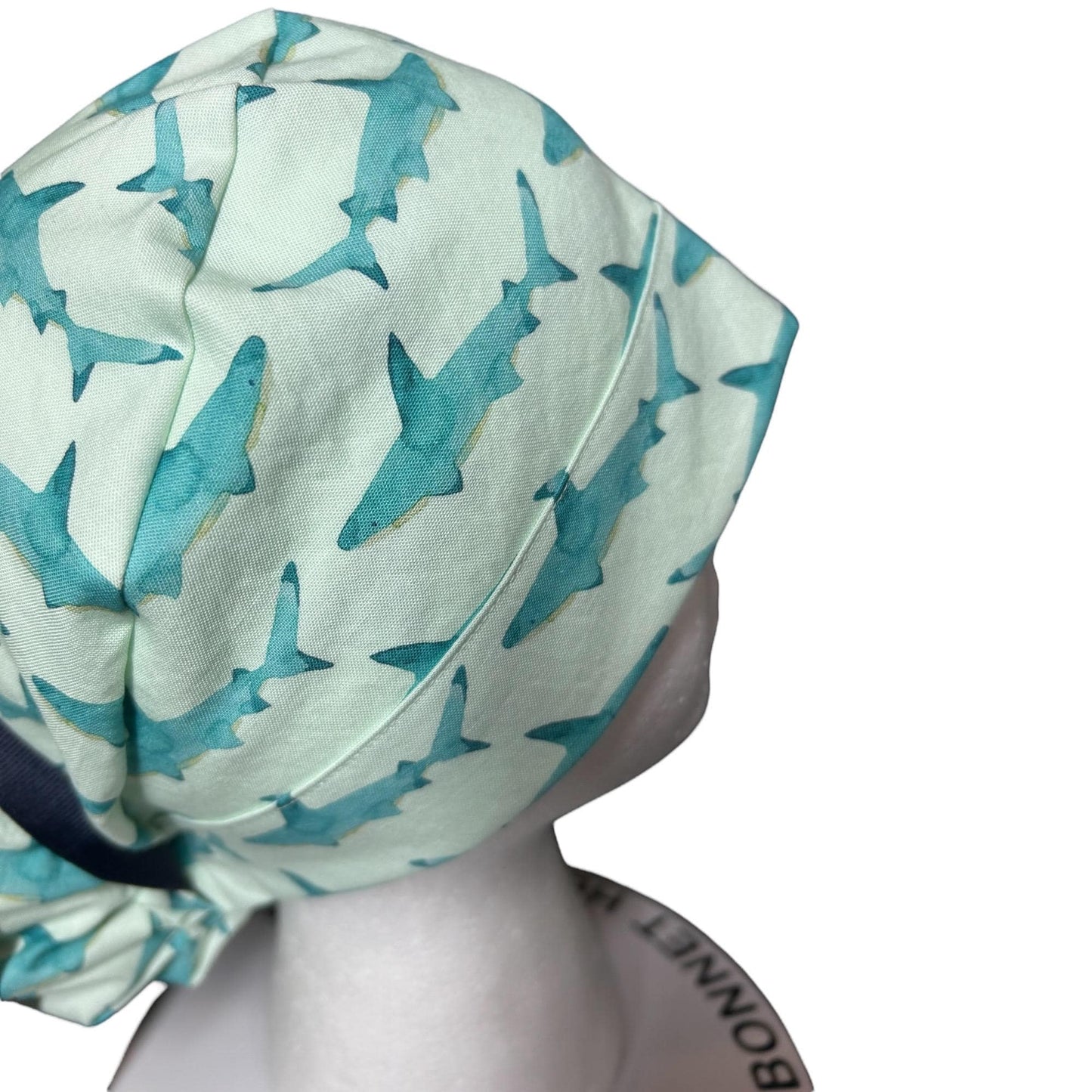 May include: A light green bonnet featuring a repeating pattern of teal sharks. The bonnet has a dark blue band and is made of soft fabric. The design is suitable for various occasions.