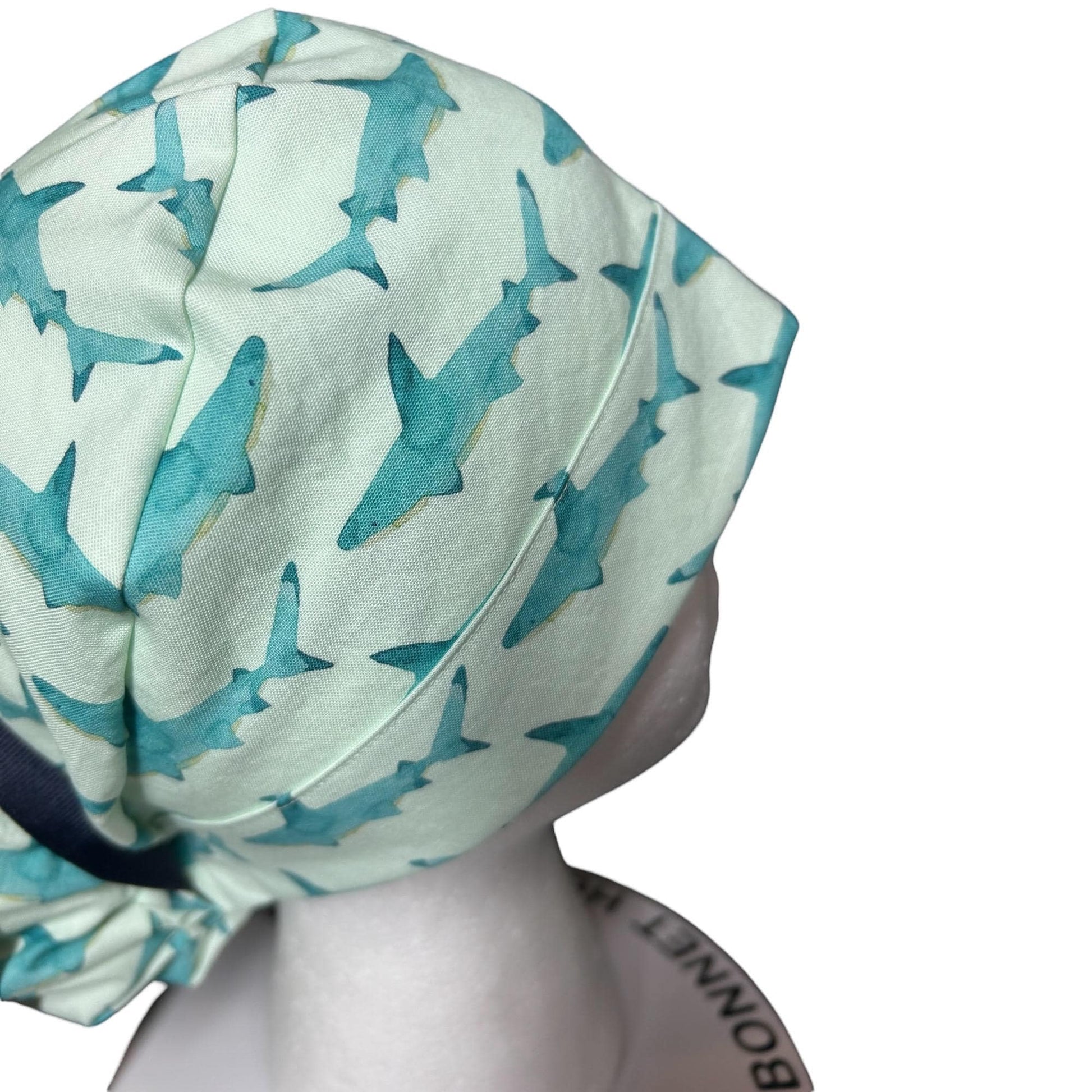 May include: A light green bonnet featuring a repeating pattern of teal sharks. The bonnet has a dark blue band and is made of soft fabric. The design is suitable for various occasions.
