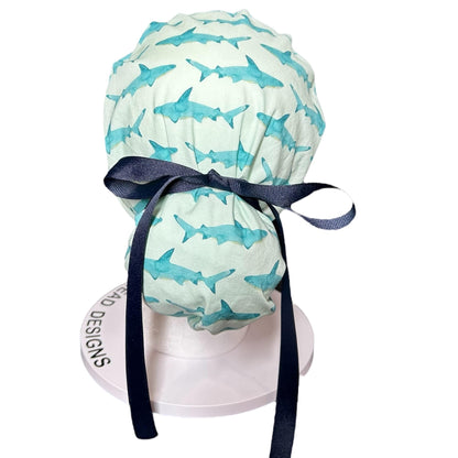 May include: A light green scrub cap featuring a pattern of blue watercolor sharks. The cap is tied with a dark blue ribbon. The cap is designed to be worn by medical professionals.