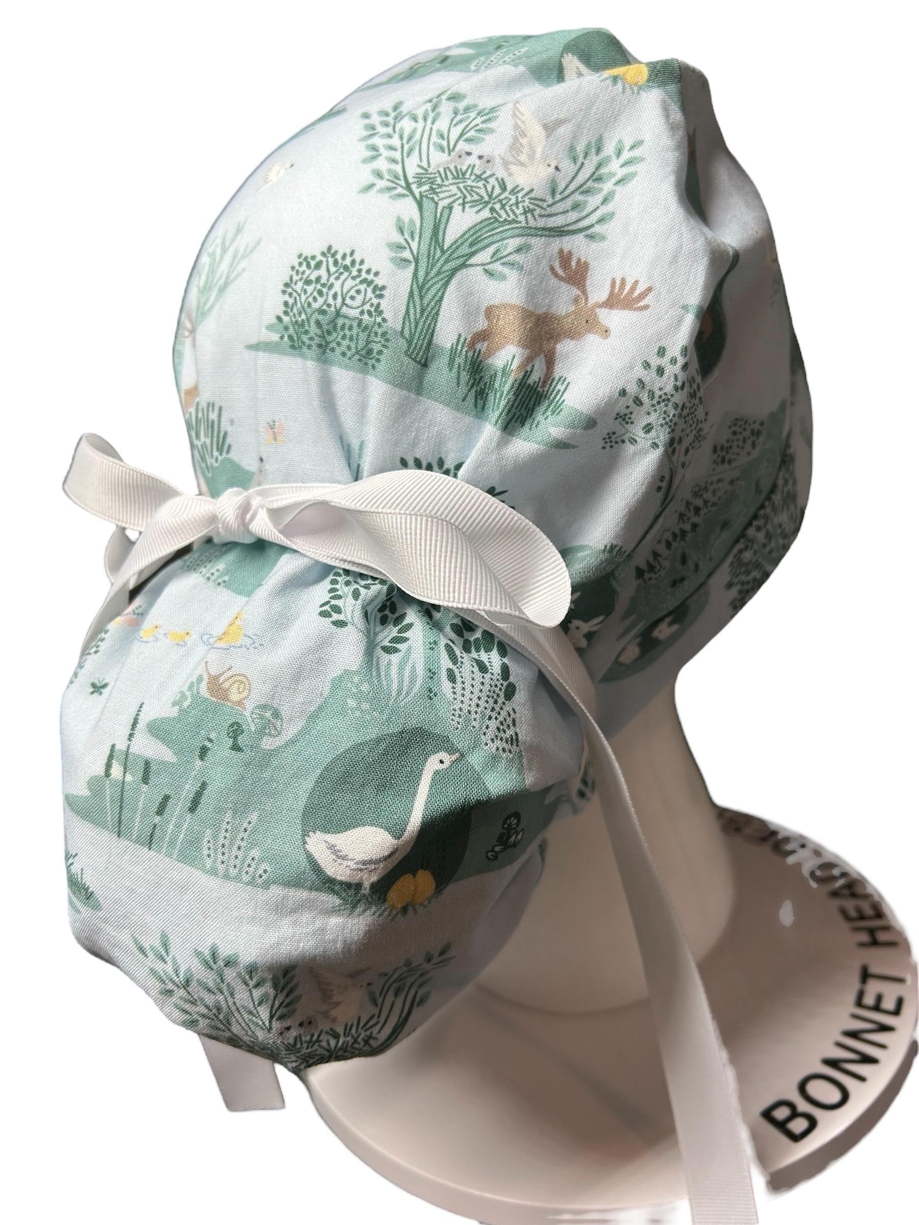 May include: A light blue scrub hat with a white ribbon tie. The hat has a woodland pattern with trees, birds, and animals in shades of green, brown, and yellow. The hat is displayed on a white mannequin head with the words 'BONNET HEAD' printed on the base.