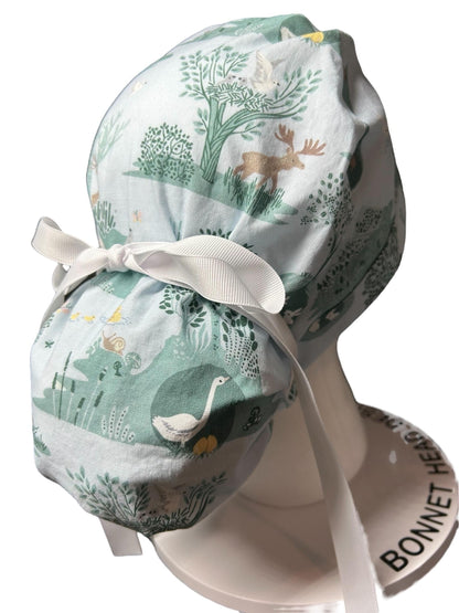 May include: A light blue scrub hat with a white ribbon tie. The hat has a woodland pattern with trees, birds, and animals in shades of green, brown, and yellow. The hat is displayed on a white mannequin head with the words 'BONNET HEAD' printed on the base.