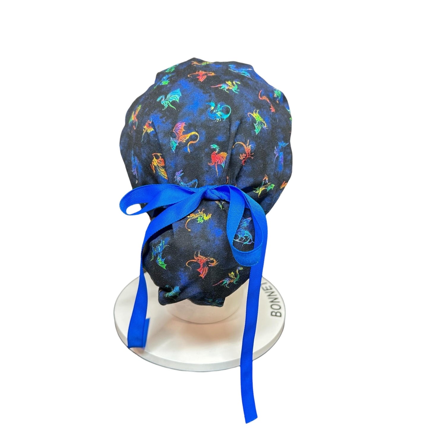 A dark blue scrub cap with a pattern of colorful dragons. The cap is tied with a bright blue ribbon. The cap is on a white stand.