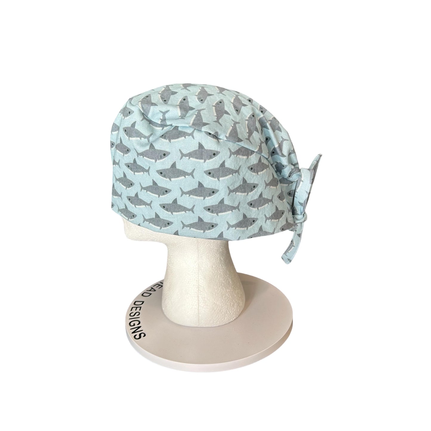 May include: A light blue surgical scrub cap with a repeating pattern of gray sharks. The cap is tied at the back and is displayed on a white mannequin head. The base of the mannequin head has the text "HEAD DESIGNS" printed on it.
