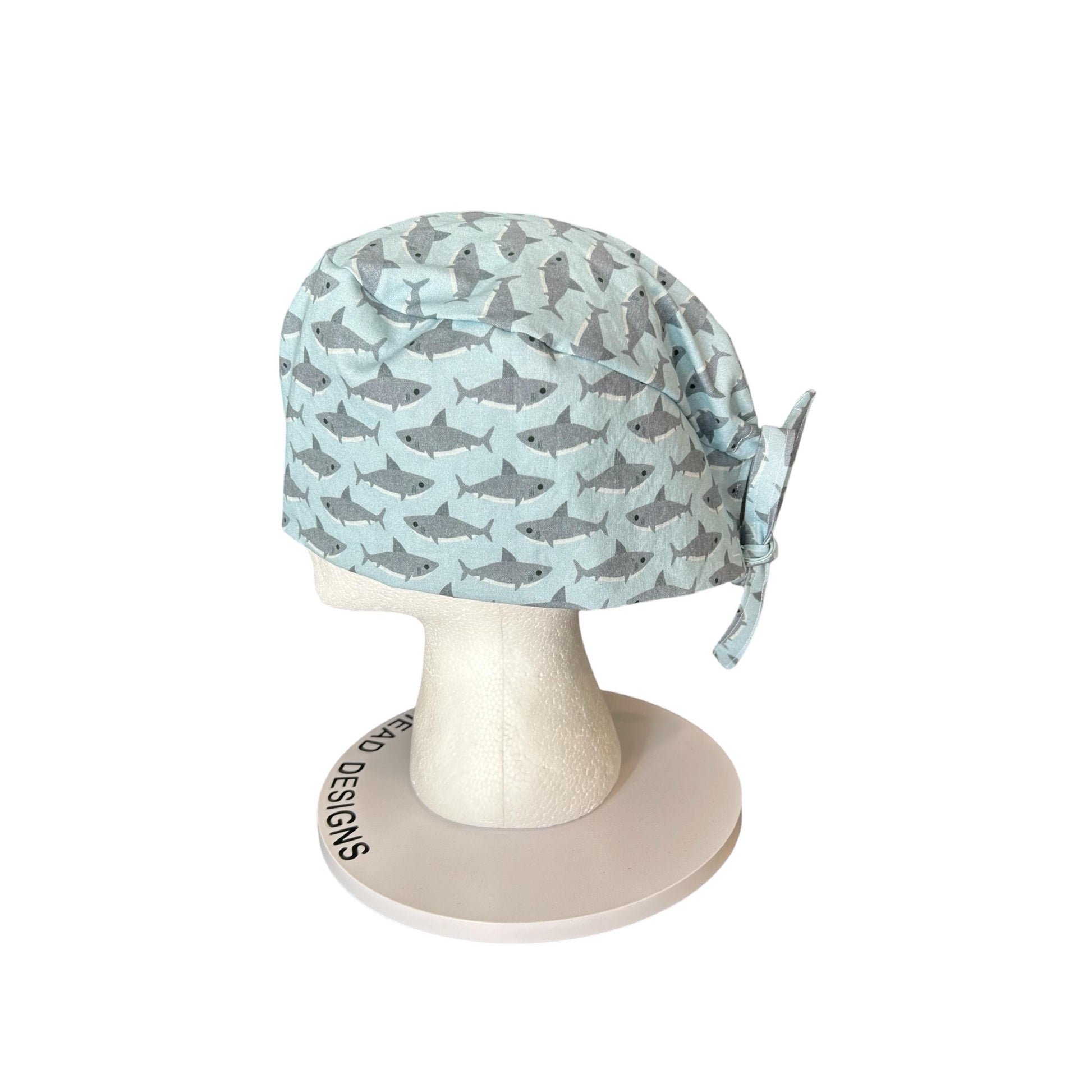 May include: A light blue surgical scrub cap with a repeating pattern of gray sharks. The cap is tied at the back and is displayed on a white mannequin head. The base of the mannequin head has the text "HEAD DESIGNS" printed on it.