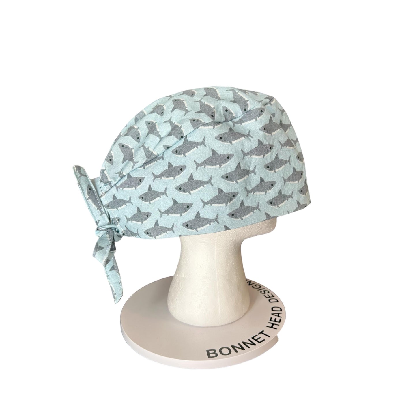 May include: A light blue surgical scrub cap with a repeating pattern of gray sharks. The cap is tied at the back with a matching fabric tie. The cap is displayed on a white mannequin head.