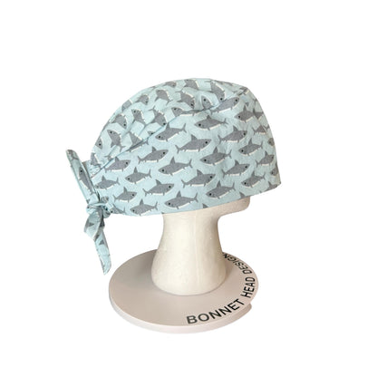 May include: A light blue surgical scrub cap with a repeating pattern of gray sharks. The cap is tied at the back with a matching fabric tie. The cap is displayed on a white mannequin head.