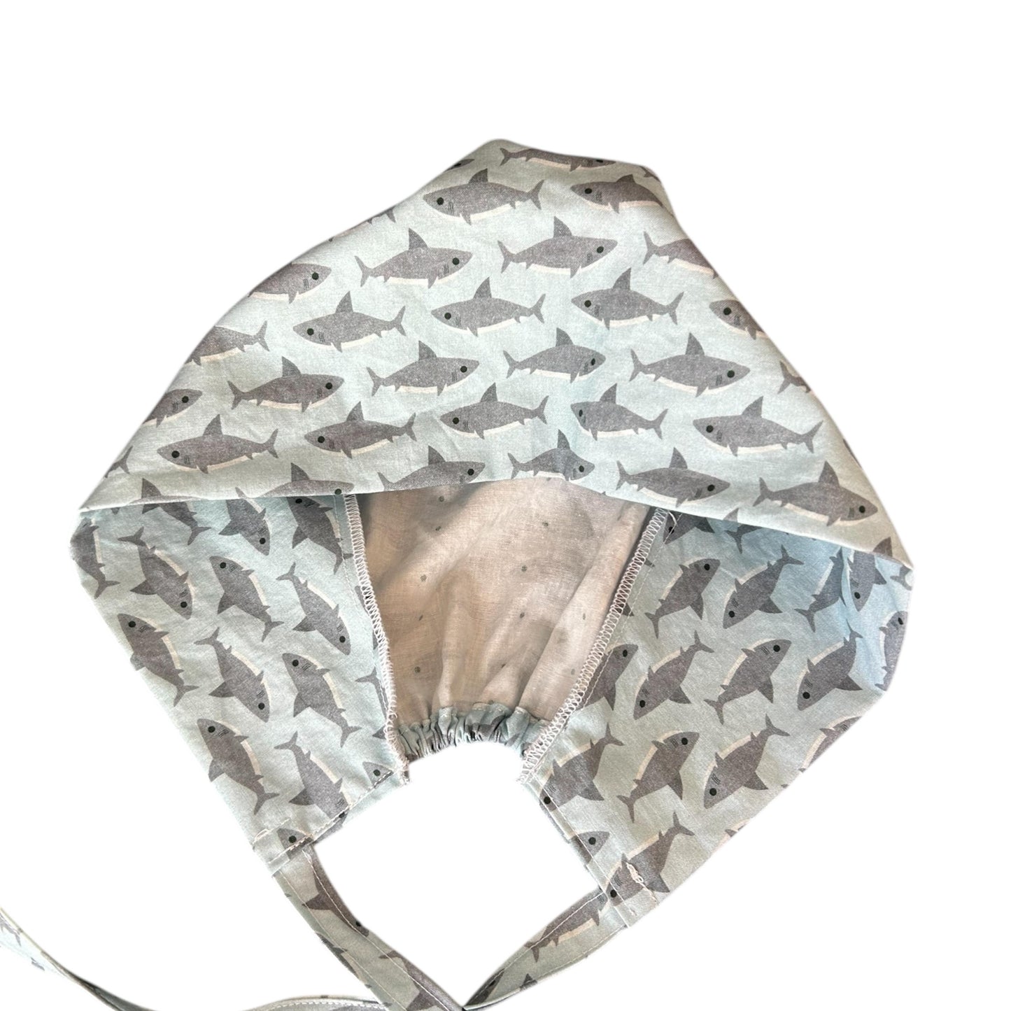 May include: A light blue surgical scrub cap with a repeating pattern of gray sharks. The cap has a gathered back and long ties for securing. The interior fabric is a light beige with small gray dots.