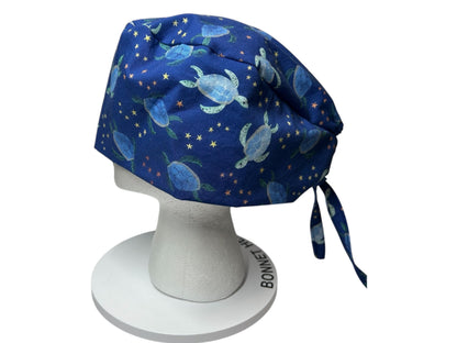 May include: A navy blue surgical scrub cap featuring a pattern of light blue sea turtles and small yellow stars. The cap is tied at the back with matching fabric ties. The cap is displayed on a white mannequin head.