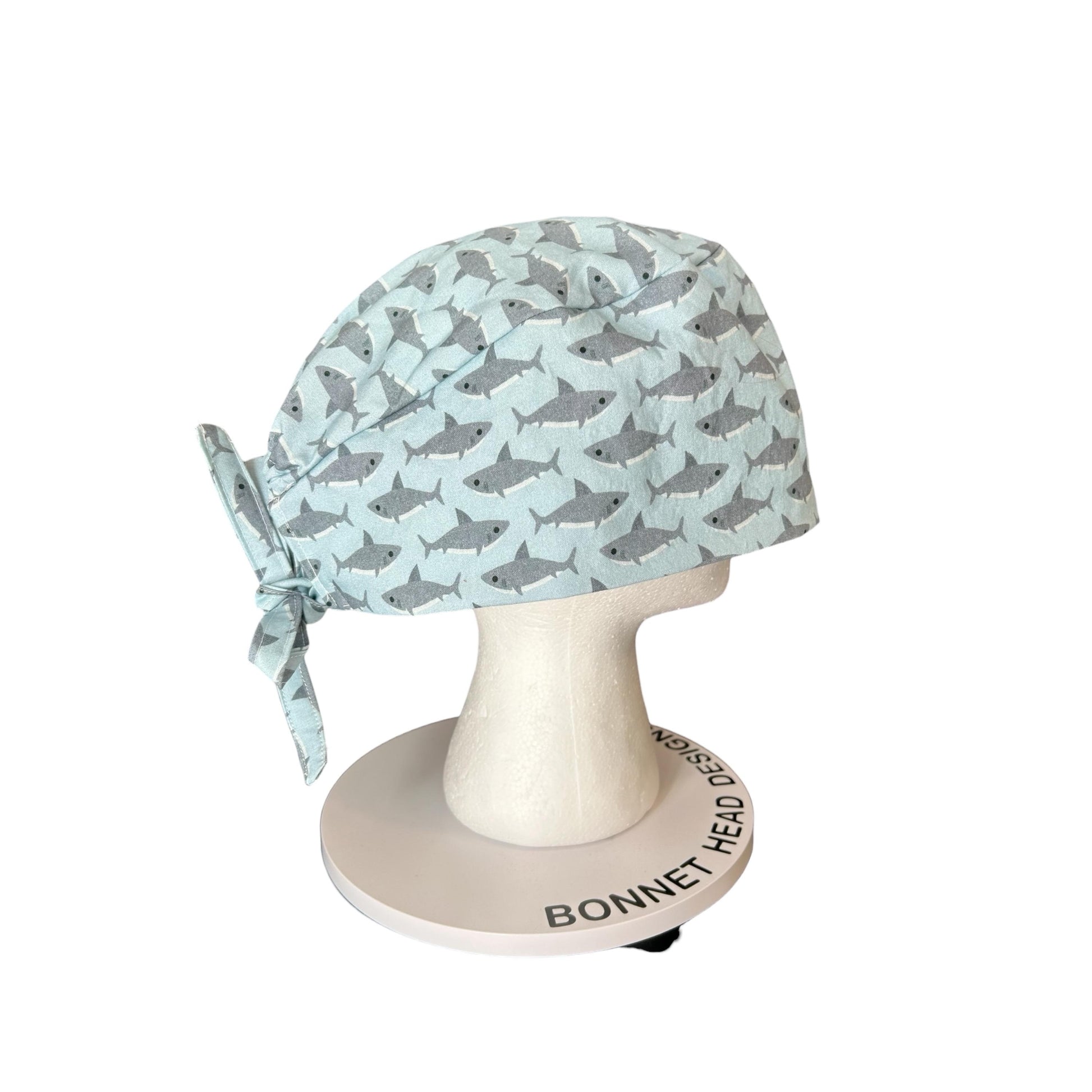 May include: A light blue surgical scrub cap with a repeating pattern of gray sharks. The cap is tied at the back and displayed on a white mannequin head. The base of the mannequin head has the text "BONNET HEAD DESIGN".