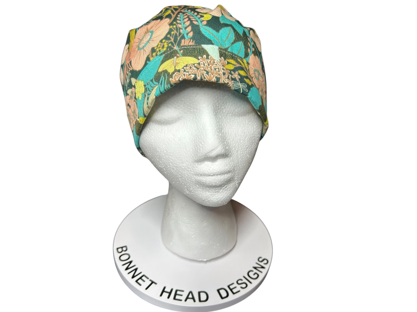 A floral print bonnet in shades of teal, peach, yellow, and green, displayed on a white mannequin head. The bonnet features a colorful botanical pattern. The base of the mannequin reads "BONNET HEAD DESIGNS".