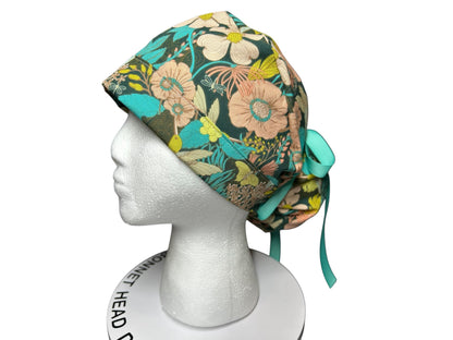 May include: A floral patterned scrub cap in shades of teal, pink, yellow, and green. The cap features a botanical design with flowers and leaves. It has a teal ribbon tie at the back. The cap is displayed on a white mannequin head.