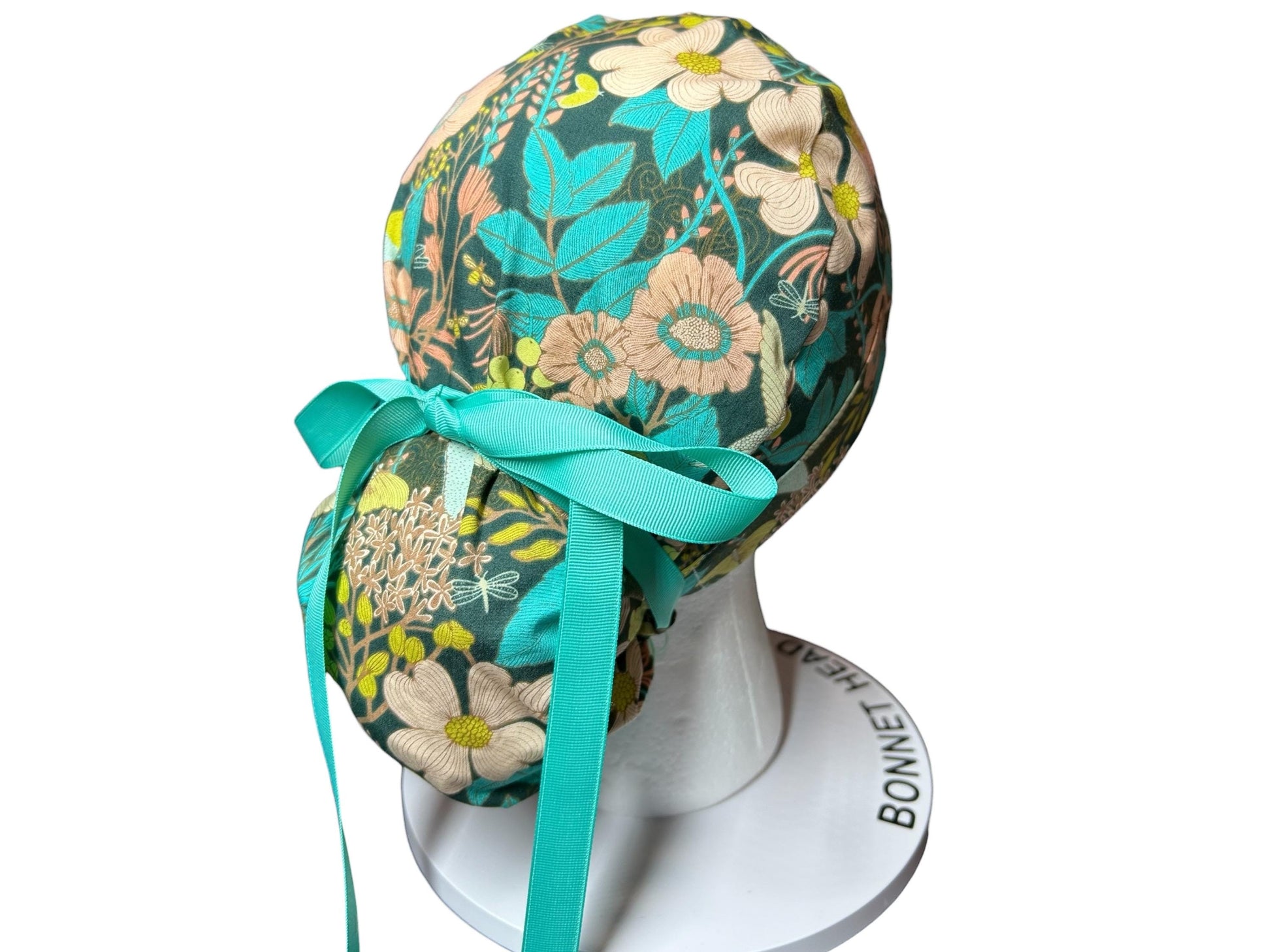 May include: A floral patterned scrub cap featuring a teal ribbon tie. The cap has a dark teal background with cream and yellow flowers, teal leaves, and a dragonfly design. The cap is displayed on a white head form.