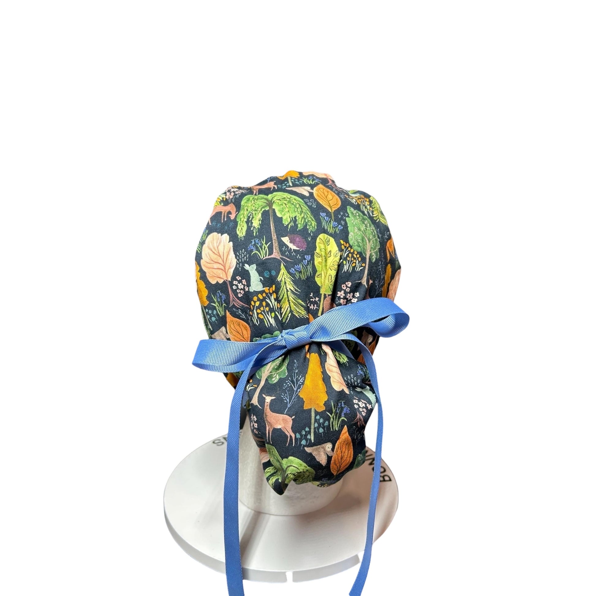 A dark blue scrub cap with a forest-themed pattern featuring trees, deer, and foxes. The cap has a light blue ribbon for tying. The cap is designed to be worn by medical professionals.
