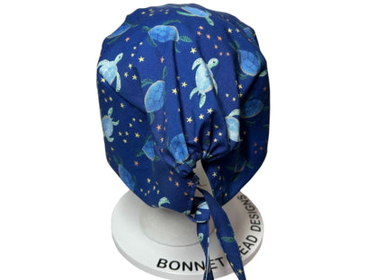 May include: A navy blue bonnet featuring a pattern of light blue sea turtles and yellow stars. The bonnet has a tie closure and is displayed on a white stand.