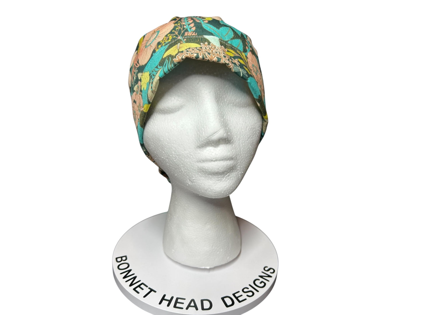 A floral print bonnet in teal, peach, and yellow hues, displayed on a white mannequin head. The bonnet features a colorful botanical pattern. The base of the display reads "Bonnet Head Designs".