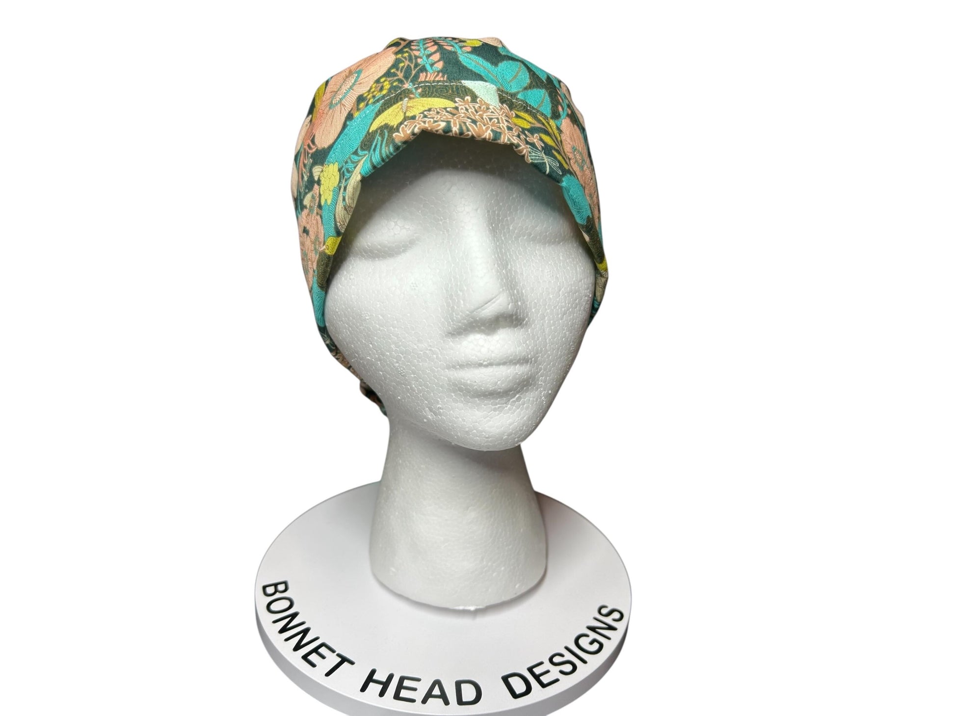 A floral print bonnet in teal, peach, and yellow hues, displayed on a white mannequin head. The bonnet features a colorful botanical pattern. The base of the display reads "Bonnet Head Designs".