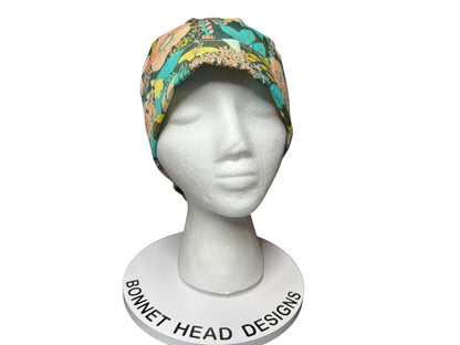 A floral print bonnet in teal, peach, and yellow hues, displayed on a white mannequin head. The bonnet features a colorful botanical pattern. The base of the display reads "Bonnet Head Designs".