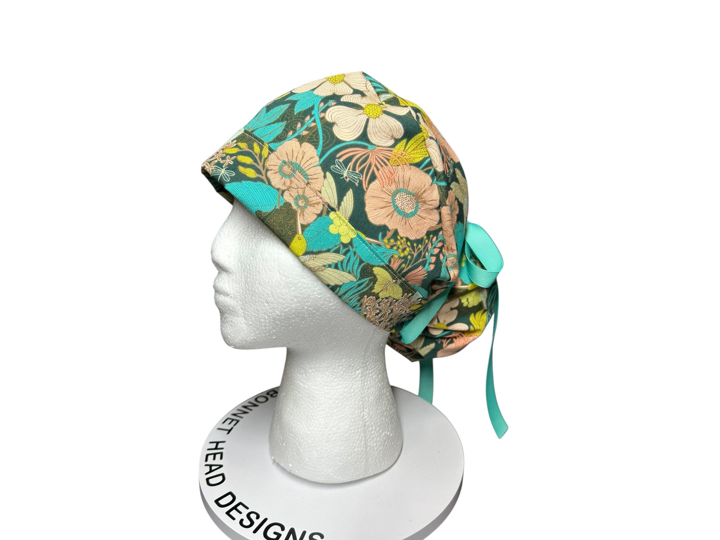 A floral patterned scrub cap with a teal ribbon tie. The cap features a mix of peach, white, and yellow flowers on a dark green background. The cap is displayed on a white mannequin head.