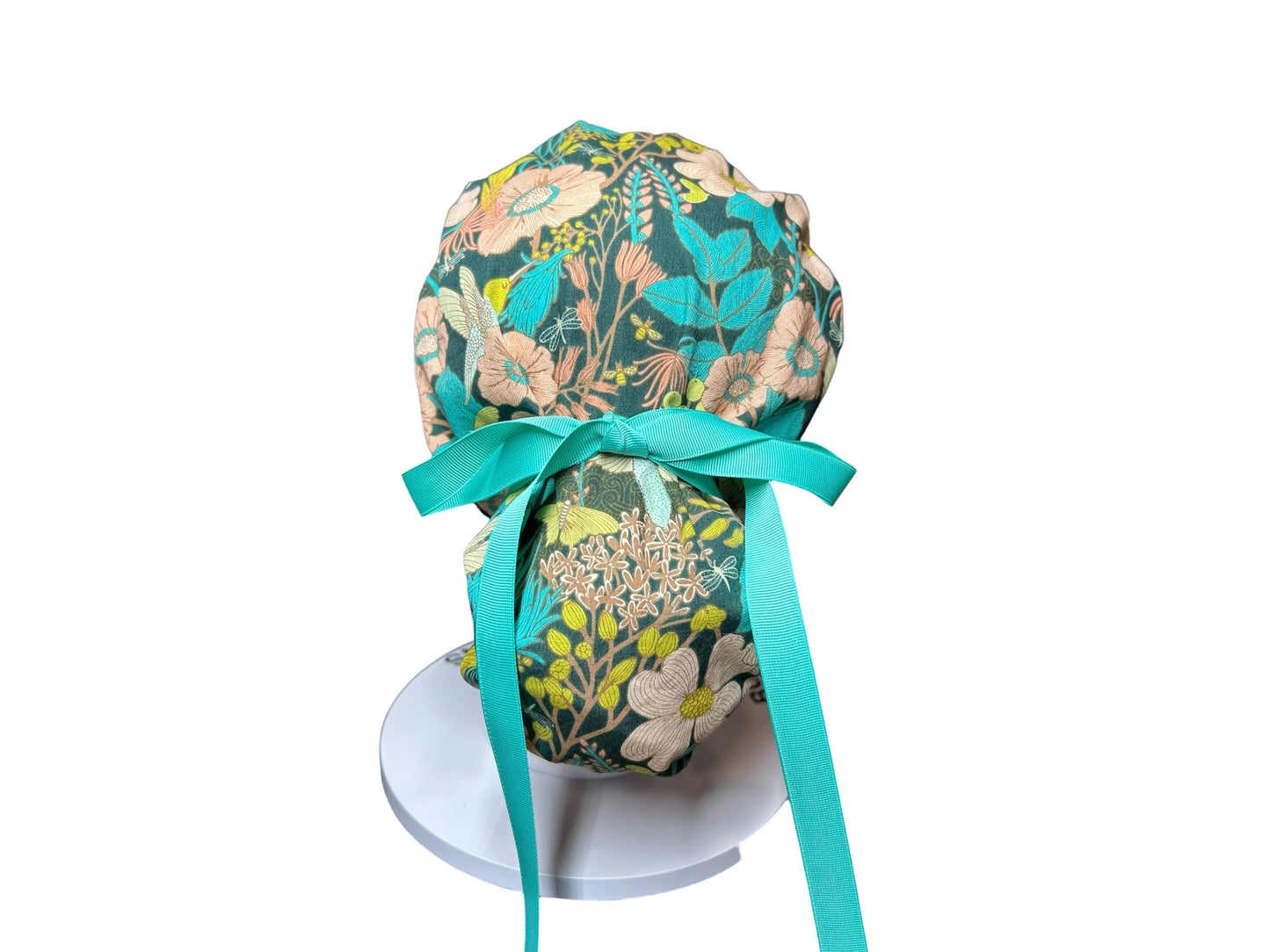 A floral patterned scrub cap in teal, peach, and cream. The cap features a teal ribbon tied in a bow at the back. The fabric design includes flowers and leaves. The cap is displayed on a white stand.
