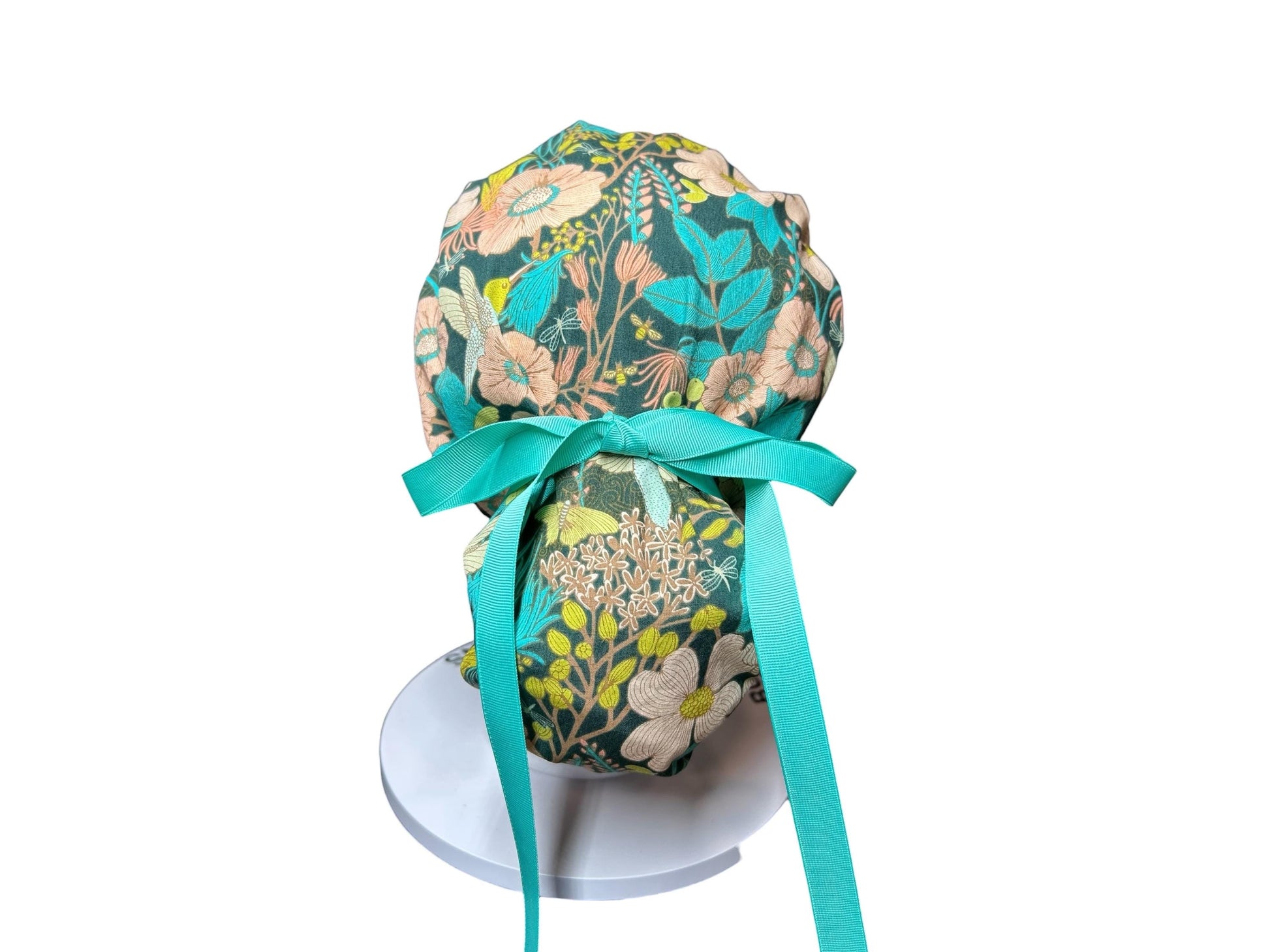 A floral patterned scrub cap in teal, peach, and cream. The cap features a teal ribbon tied in a bow at the back. The fabric design includes flowers and leaves. The cap is displayed on a white stand.