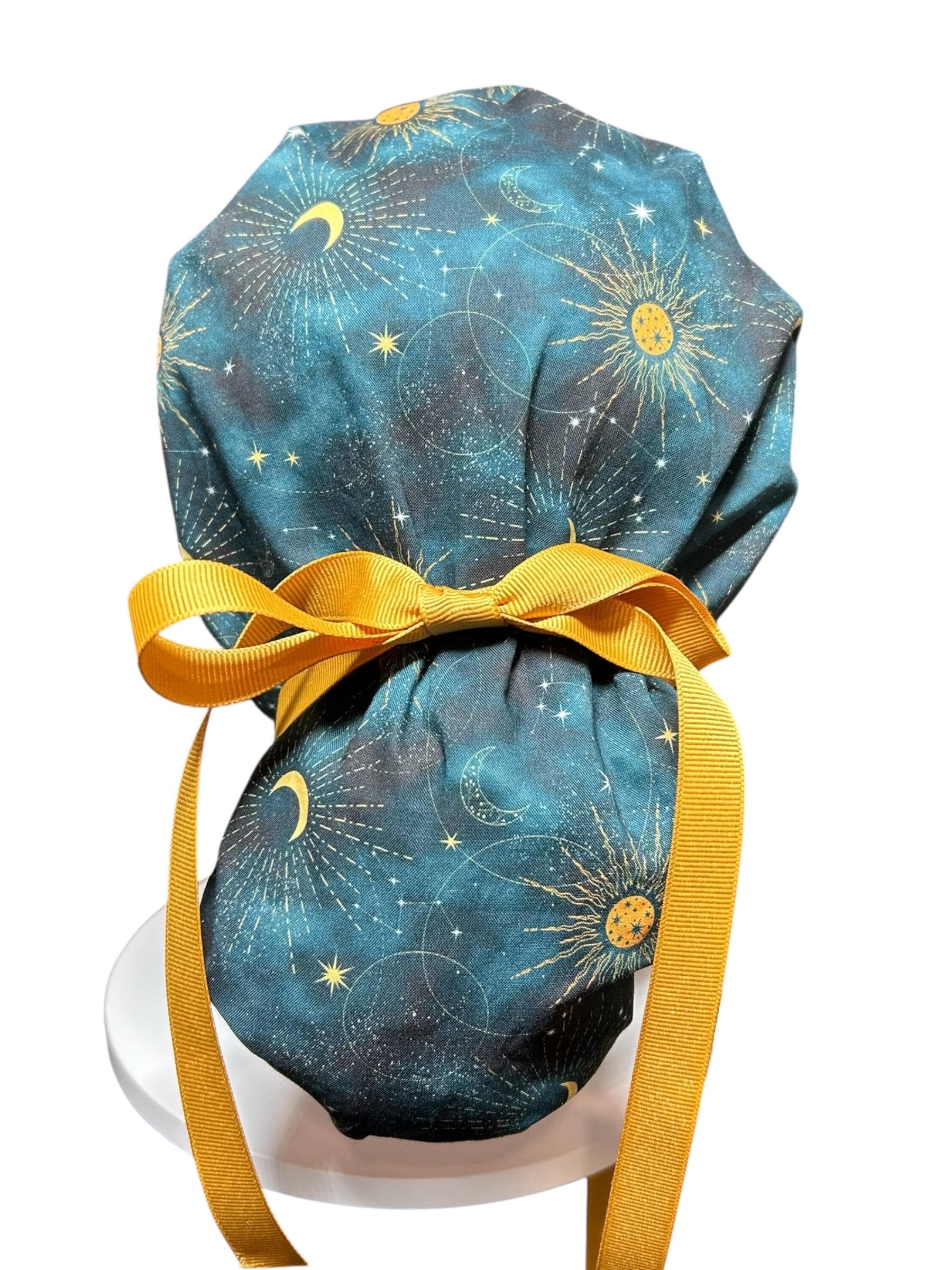 A teal and gold celestial print scrub cap with a mustard yellow bow and ties. The fabric features a pattern of suns, moons, and stars. The cap is designed to be worn in a medical or professional setting.