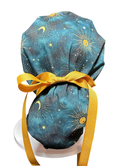 A teal and gold celestial print scrub cap with a mustard yellow bow and ties. The fabric features a pattern of suns, moons, and stars. The cap is designed to be worn in a medical or professional setting.