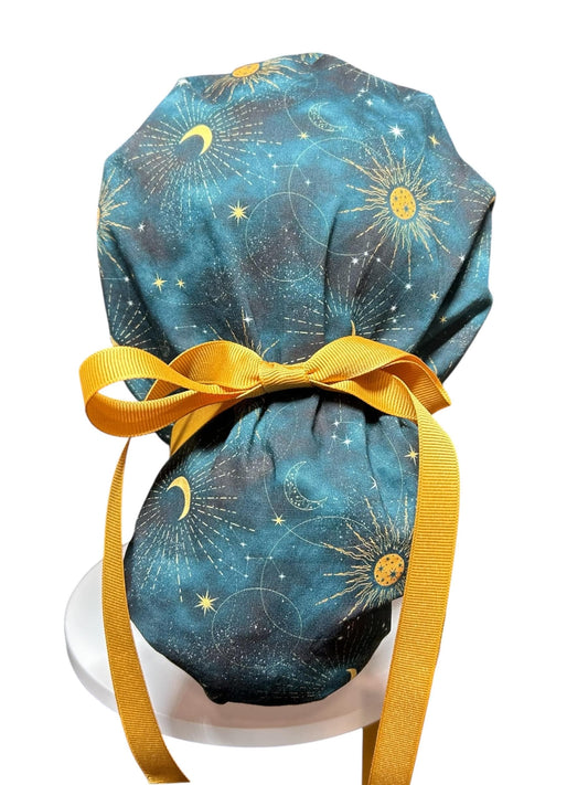 A teal and gold celestial print scrub cap with a mustard yellow bow and ties. The fabric features a pattern of suns, moons, and stars. The cap is designed to be worn in a medical or professional setting.