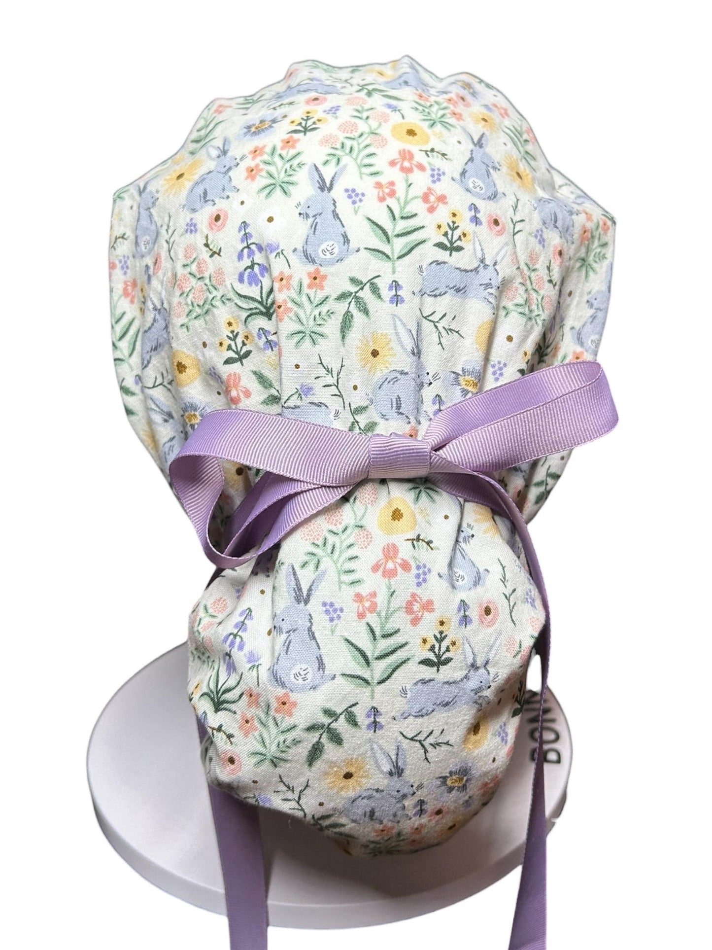 A white scrub cap with a floral and bunny print. The cap features a light purple ribbon bow and ties. The fabric design includes pastel flowers and light blue bunnies, creating a spring or Easter theme.