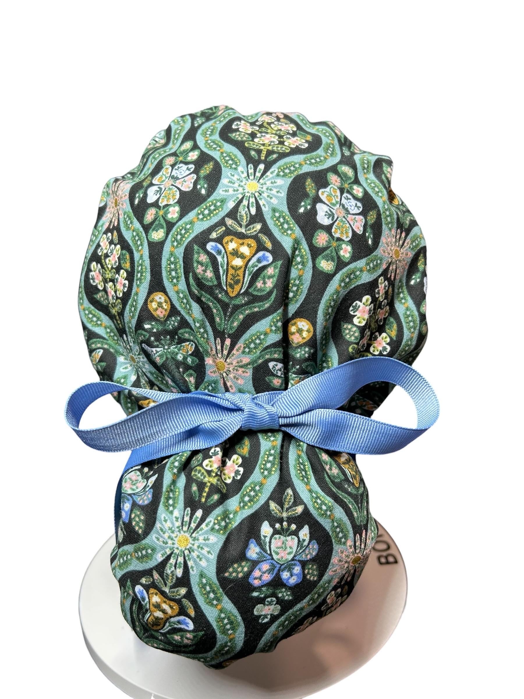 A patterned scrub cap featuring a floral design in black, teal, and pink. The cap is tied with a light blue ribbon. The fabric has a repeating pattern of flowers and leaves.