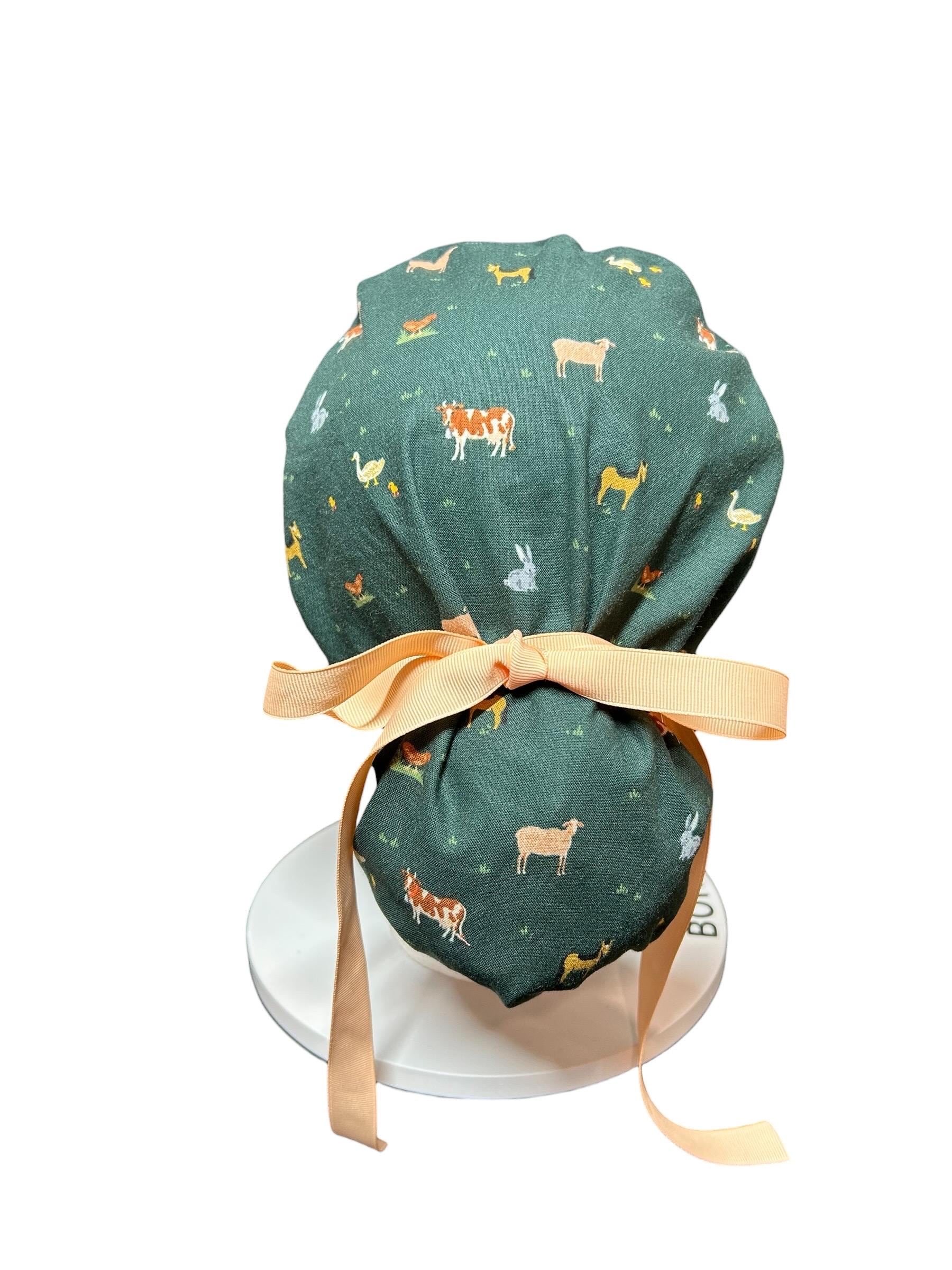 A dark green scrub hat with a farm animal pattern, including cows, sheep, chickens, and rabbits. The hat is tied with a peach-colored ribbon. The hat is designed to be worn in a medical or veterinary setting.